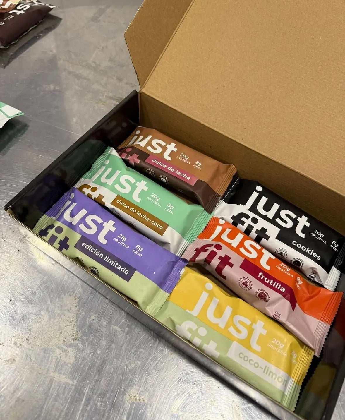Una caja abierta con barras Just Fit de seis sabores: dulce de leche, dulce de leche y coco, cookies, frutilla, coco-limón y edición limitada.
